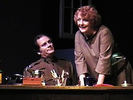 A scene from a stage play with two older actors behind a desk. One actor is seated and attired in a World War I uniform, and the other actor is standing and wears a brown dress.