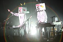 Two people with white comedy space suit costumes ant include a square tv sort of masks on stage in front of instruments and microphones