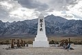 Photograph of a memorial at the Manzanar War Relocation Center. A low white obelisk with Japanese kenji characters and flower offerings stands against an expansive backdrop of the Owens Valley floor, snowy mountains, large clouds, and blue sky.