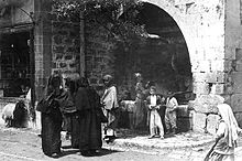 From left to right: A boy staring out from a store's window sill beneath which a lamb is walking by; three fully veiled women conversing on the street; beneath an olive grove jutting out of a large stone archway and beside a fountain, a man is walking, a woman is collecting water from the fountain, and two young boys are standing and smiling; a young girl walking on the street