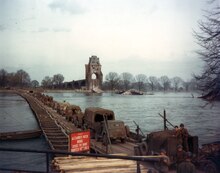 Trucks cross the bridge. The ruins of the original bridge are in the background. A sign says: "Alexander Patch bridge – cross the Rhine courtesy of the 85th Engineers"