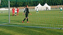 Von hinten durch ein Fußballtor fotografiert, sieht man die Torhüterin der koreanischen Frauenmannschaft im schwarzen Trikot von hinten. Auf dem Spielfeld mehrere koreanische Spielerinnen in Rot und Spielerinnen der Elfenbeinküste in weißem Shirt und grüner kurzer Hose, im Hintergrund weiße Zelte.