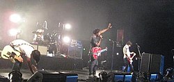 Two guitarists, a drummer, and a bassist are on a stage lit by white lights. One of the guitarists is kneeling to change his pedals and several speakers are visible in the foreground.