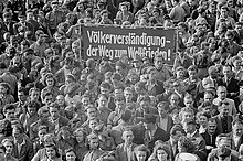 "Schwarz-Weiß-Fotografie zeigt Menschenmenge und ein Plakat mit der Aufschrift: Völkerverständigung – Der Weg zum Weltfriedenǃ"