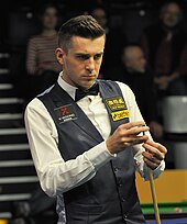 Mark Selby stands looking at a table while he chalks the cue in his left hand.