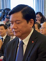 Đinh La Thăng looking down wearing a black suit, red tie and a National Assembly of Vietnam pin on his right chest.