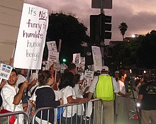 A crowd of people at the left are standing behind a gated barrier. The crowd is holding protest signs and the majority are looking away from the camera. An obscured man is being interviewed at the right side of the image. The sky appears to be approaching night.