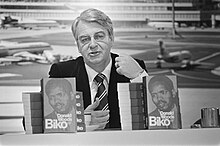A black and white photograph of a middle-aged white man. He has grey hair and is wearing a black suit and tie. He is sitting at a table, and in front of him are two upright books; both feature the face of Steve Biko, a young black man.