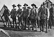 Men in military uniforms with rifles and bandoliers stand at ease in ranks