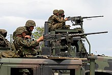 In the foreground, a HMMWV, with a MTVR in the background. Both vehicles have M2 machine guns mounted and U.S. Marines firing them.