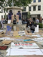 The pedestal is seen with purple spraypaint graffiti "BLM" over two of the bronze plaques and black "Black Lives Matter" and stencilled raised fists on the plinth. Placards propped on the pedestal include "Black Lives Matter", "Silence is Violence", "The UK is not innocent" and "In unity is strength". Many more placards lie on the ground around the pedestal, with "Black Lives Matter","Racism is a global pandemic" and other slogans.