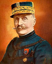 A gentleman with a handlebar-style moustache. He is wearing a peaked cap highly decorated with gold braiding. He wears a dark blue, high-collared jacket, with regularly spaced horizontal bands of dark braiding. On his left breast, he wears a medal attached to a red ribbon.