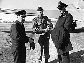 Three-quarter informal portrait of three men in military uniforms in front of the left wing of a twin-engined aircraft