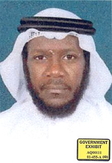 Mustafa al-Hawsawi sitting crosslegged on a green prayer rug. He is wearing a brown pakol hat and a white T-shirt. He is looking directly into the camera.