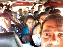 A middle-aged white man takes a selfie inside a very taxi brousse. There are at least 17 passengers behind him, ranging from infant to later age; some are making unpleasant expressions.