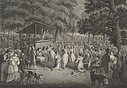 A black-and-white image depicting a Methodist camp meeting during the Second Great Awakening. A preacher, his arms raised up, sermonizes on a small wooden stage with a canopy over him. The audience is a huge crowd of people, mostly standing, though a few sit on benches. Some seem to talk to each other, some seem to listen raptly. One man kneels, as if praying. One woman has swooned and fallen over and is held up a man. There is a mix of dress, with some people in very fine clothing. The setting is apparently a forest, as large trees frame the image. In the background, dozens of tents are visible.