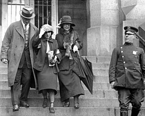A black and white photograph of Jazz Age bandit Celia Cooney leaving a New York courthouse. Attired in a dark suit with a Cloche hat, Cooney is escorted on one side by a plainclothes policeman and a female warden on the other. On the far right of the photograph, a New York City policeman stands in full uniform.