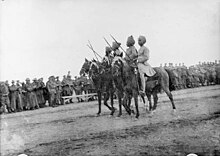 In a black and white photograph, four men with swords raised at 45 degrees wearing military uniforms and turbans ride on dark-coloured horses facing left. Behind the men, a large crowd of soldiers look on.