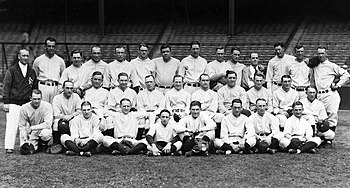 A posed team photo of a baseball team. They wear uniforms and are seated and standing in three rows, with a man in a jacket standing to the left.