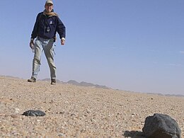 Low angle picture from ground showing sand and a large rock with a man looming a few feet back against a blue sky.