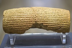 Rear view of a barrel-shaped clay cylinder resting on a stand. The cylinder is round with square edge very close up. It is covered with lines of cuneiform and has a hollow centre. The centre is lined with plate measuring around 10 millimeters in thickness.text