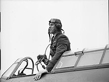 A black and white photograph of a man standing in the cockpit of an aircraft; he wears a flying helmet