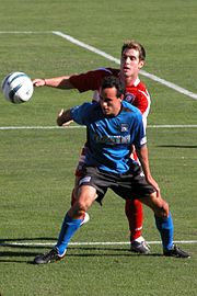 Landon Donovan, wearing a blue jersey, uses his body to hold off a defender wearing a red jersey.