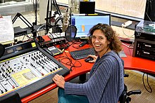 Lächelnde Frau vor einem Mischpult und Mikrofon, am rechten Bildrand ist ein Computerbildschirm