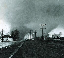 Two greyish tornado funnels silhouetted against a pale sky on either side of a road behind telephone poles