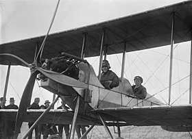 Two men in flying gear seated in tandem open cockpits of a biplane with a four-bladed propeller