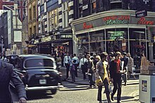 Colour photo of a busy city intersection with two young white males walking across in the foreground