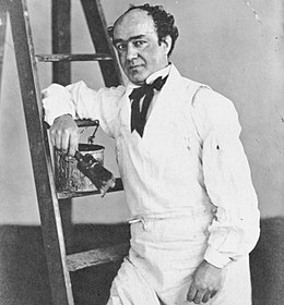 Oxford, leaning against a ladder, holding a paintbrush. He has black, curling hair, the has receded to the top of his head; he is of medium build.