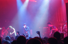 Two guitarists, a drummer, a bassist, and a vocalist are performing a song live on a stage lit by red and blue lights. The crowd, some of whom have their hands in the air, are visible in the foreground.