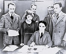 Frontale Schwarzweißfotografie von einem Mann, der an einem Tisch mit mehreren Papieren und Stiften sitzt. Er blickt in die Kamera und hält in seiner rechten Hand einen Füllfederhalter. Hinter ihm stehen vier Männer und eine Frau, die ihn anschauen. Der linke Mann hält Papier in seinen Händen.