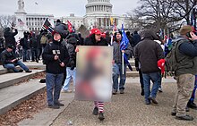 A Trump supporter carries a QAnon-tagged placard with Jesus wearing a MAGA hat at the moment the U.S. Congress was violently attacked by rioters on January 6, 2021. (The placard is blurred for reasons of copyright.)