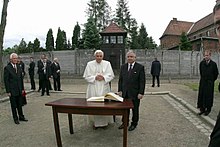 Farbige Fischaugenperspektive vom Papst in weißem Papstgewand und einem Mann in dunklem Anzug, die vor einem Holztisch mit einem aufgeschlagenen Buch stehen. Um sie herum stehen mehrere Männer in dunkler Kleidung. Sie stehen in einem Hof, umringt von einer Betonmauer und einem Wachturm.