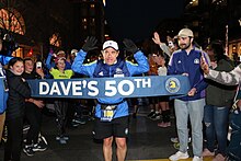 A man in a white hat, blue jacket, and black shorts breaks a finisher tape that says "Dave's 50th" and shows the Boston Marathon logo, surrounded by a crowd of people cheering