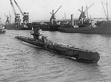 A surfaced submarine is slowly sailing into a port. A wharf can be seen in the background with large cranes, buildings and two berthed ships.
