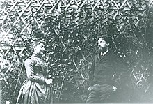 Schwarz-Weiß-Fotografie von zwei Personen vor einem Gitter mit Kletterpflanze. Die Frau auf der linken Seite trägt ein Kleid und hochgesteckte Haare. Der Mann auf der rechten Seite Vollbart und einen Anzug. Sie sind einander zugewandt, halten aber Abstand. Die Frau lächelt.
