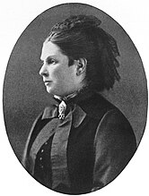 Black-and-white photographic portrait of a standing woman, formally dressed, facing left
