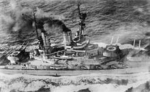 Overhead view of a large battleship; black smoke pours from its smoke stacks as it steams through choppy seas.