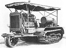 An early bulldozer-like tractor, on crawler tracks, with a leading single wheel for steering - projecting from the front - on an extension to the frame. The large internal combustion engine is in full view, with the cooling radiator prominent at the front. An overall roof is supported by thin rods, and side protection sheeting is rolled up under the edge of the roof.