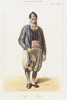 A bearded man wearing a black hat and vest and white large baggy trousers holding a blue cape on his shoulder