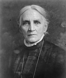 An older white woman with white hair, wearing a high-collared dark dress with lace trim