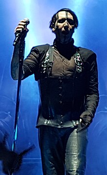 Image of the band's vocalist wearing black clothes and holding a microphone in his right hand. The background of the image is illuminated with blue stage lighting, and on the bottom left corner the hand of a concert-goer can be seen giving the 'devil-horns' signal.