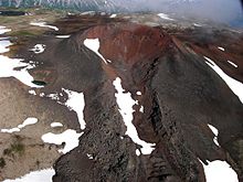 A dark-coloured volcanic cone with a summit crater rising above a sparsely snow-covered rocky plateau.