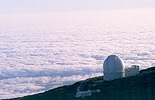 Aerial view of white domed building on side of mountain with floor of white clouds extending to the horizon below and behind the mountain.