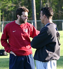 Ben Olsen, wearing a red jersey, converses with an unknown man at a soccer field.