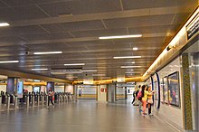 A large bright space with tiled floor and suspended ceiling with occasional lights. A row of ticket gates are on the left