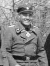 A smiling man wearing a military uniform, peaked cap with skull emblem and neck order in the shape of a cross.